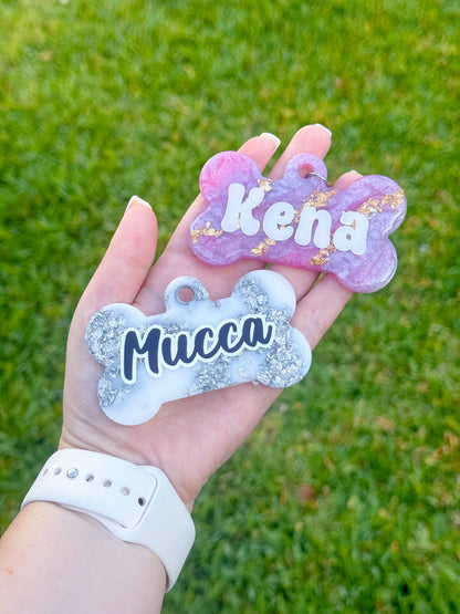 Placas en forma de hueso con glitter en colores blanco y morado con nombres Mucca y Kena. Diseño brillante y duradero