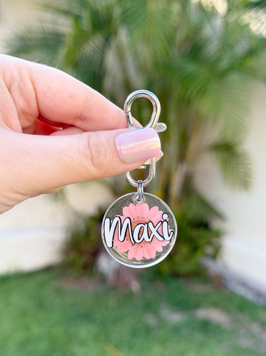Placa redonda con flor prensada y nombre “Maxi”. Diseño delicado en tono rosado con gancho metálico. Ideal para perritos con estilo.