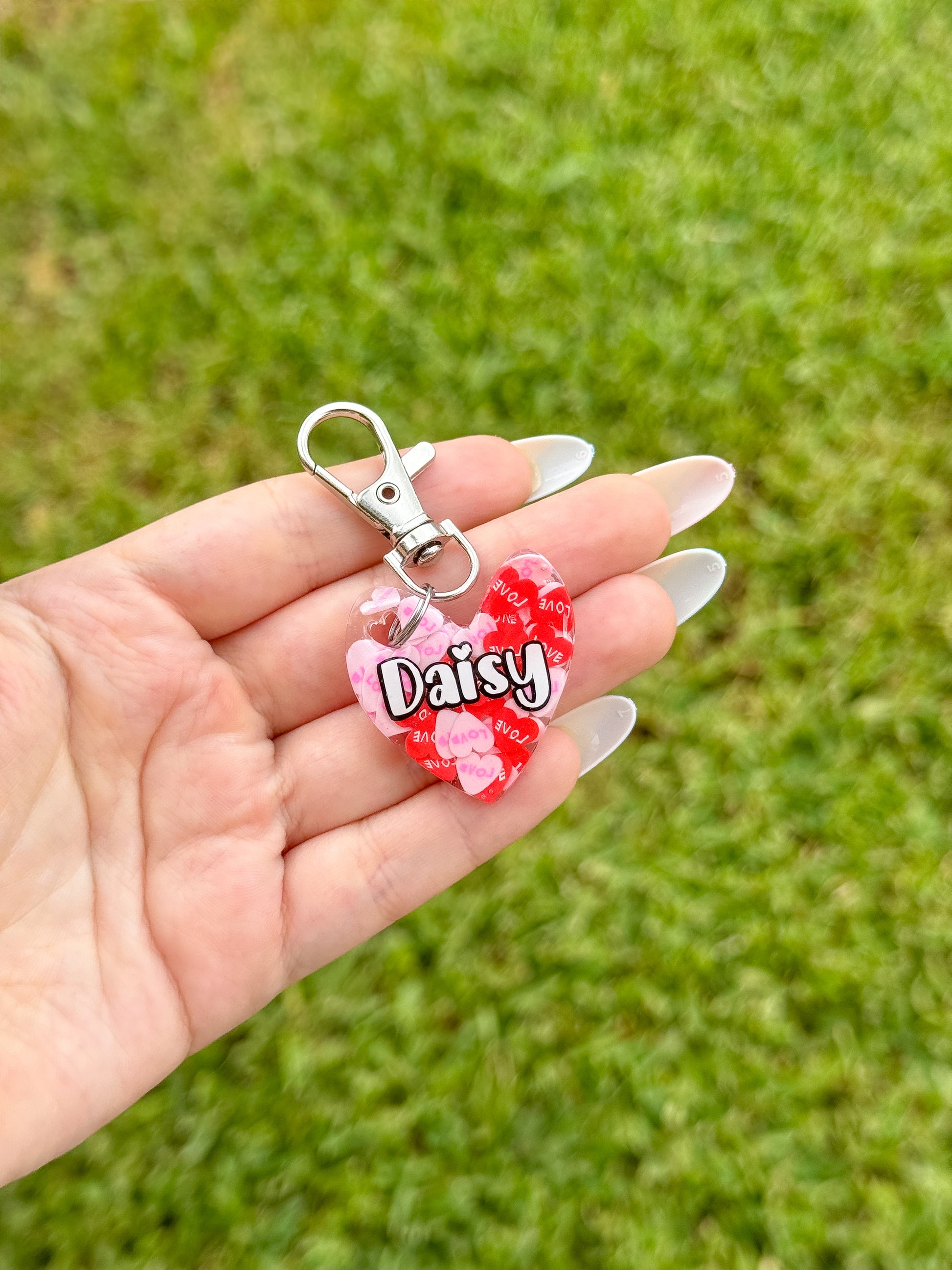 Placa roja en forma de corazón con pompón y broche, personalizada con el nombre “Daisy